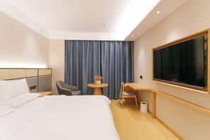 Une chambre d’hôtel avec un grand lit, un bureau, une chaise, une petite table avec un vase et une grande fenêtre avec des rideaux.