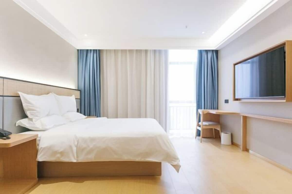 A modern hotel room with a large bed, a bedside table, a TV mounted on the wall, and a balcony with curtains.