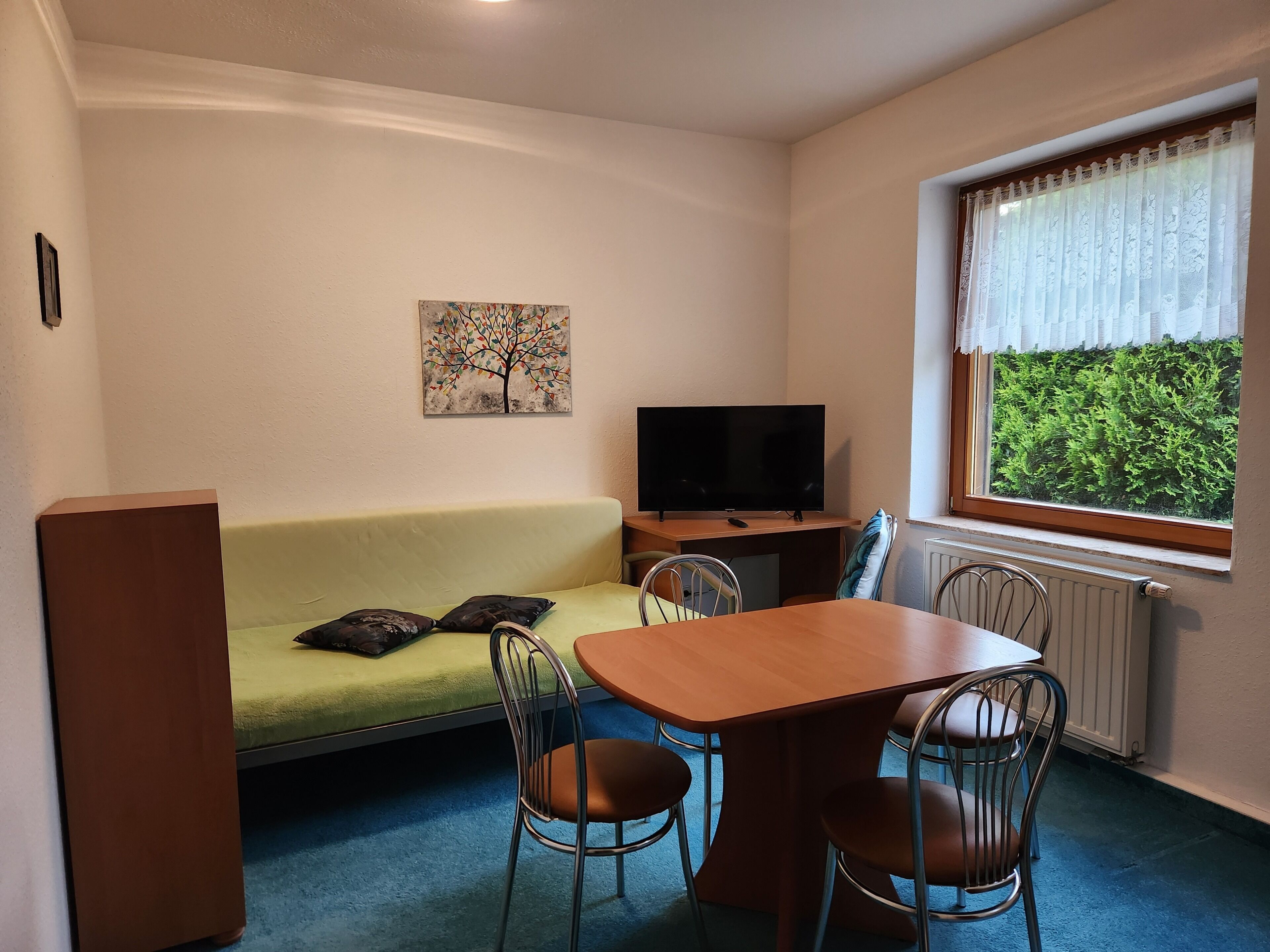 Een kamer met een groene bank, een houten tafel met stoelen, een televisie en een raam met uitzicht op groen.