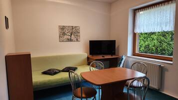 Een kamer met een groene bank, een houten tafel met stoelen, een televisie en een raam met uitzicht op groen.