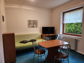 A room with a green sofa, a wooden table with chairs, a television, and a window with a view of greenery. - Siegmundsburger Werraquelle House, "Rennweg" Apartment—first floor, pets allowed (Siegmundsburg)