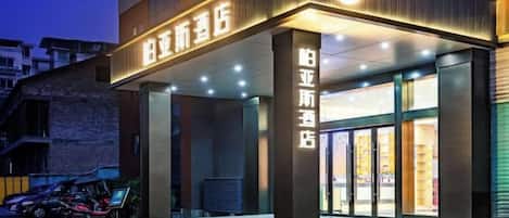 A hotel entrance with a modern design, featuring a prominent illuminated sign, a glass facade, and a visible entrance area with steps leading up to the door.