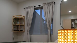 A bedroom with a bed, a bedside lamp, a window with curtains, a shelf with a plant, and a mirror.
