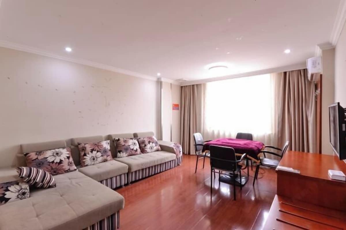 A living room with a sofa set, a dining table with chairs, a wooden desk, and a large window with curtains.