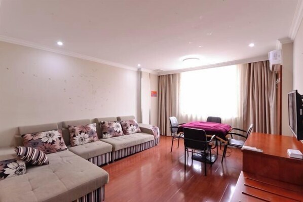 A living room with a sofa set, a dining table with chairs, a wooden desk, and a large window with curtains.