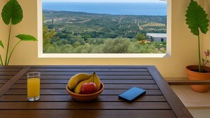 Une table avec un saladier de fruits, un verre de jus et un smartphone, avec vue sur un paysage côtier à travers une fenêtre en arc de cercle.