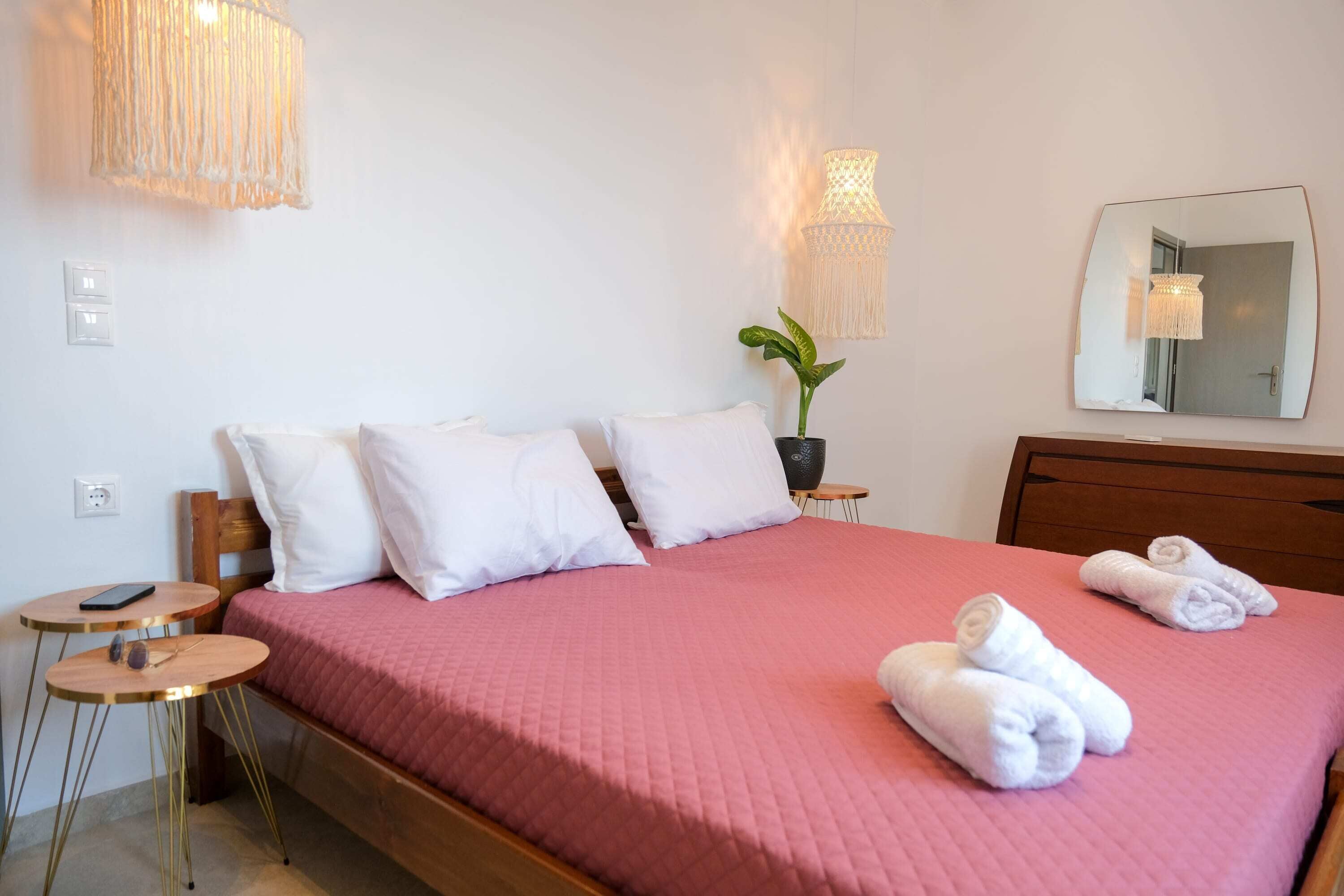 A neatly made bed with white pillows and a pink bedspread, a small round table with a phone and keys, a potted plant, and a mirror on the wall.