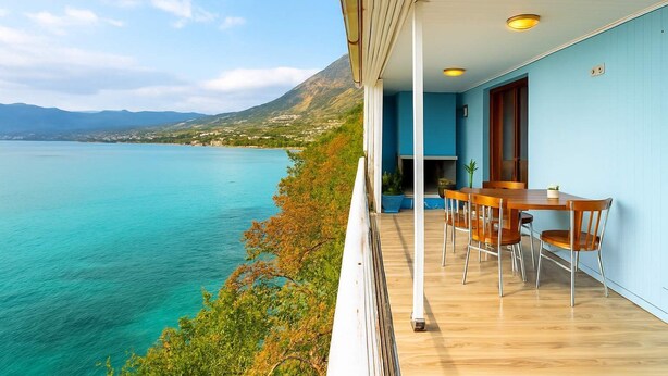 A balcony with a wooden table and chairs, overlooking a turquoise sea and a mountainous landscape. - Endless Blue Seafront Retreat - Mikra Mantineia (Paralia Vergas)