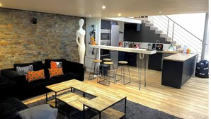 A modern living room with a black sofa, wooden coffee table, and a stone wall. A kitchen with a bar and stools is visible in the background. - Loft Proche Centre Paris y Loger ou Professionnel (Suresnes)