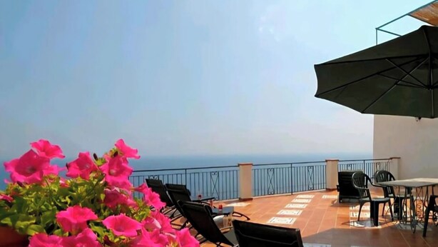 A terrace with chairs and tables, a potted plant with pink flowers, and a view of the sea. - TERRAZZA DI ROSA-SEA VIEW (Praiano)
