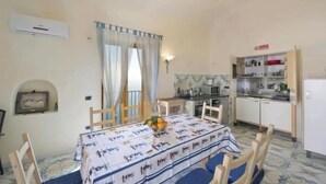 A dining area with a table set for four, a kitchen with open cabinets, and a small alcove with a picture. - TERRAZZA DI ROSA-SEA VIEW (Praiano)