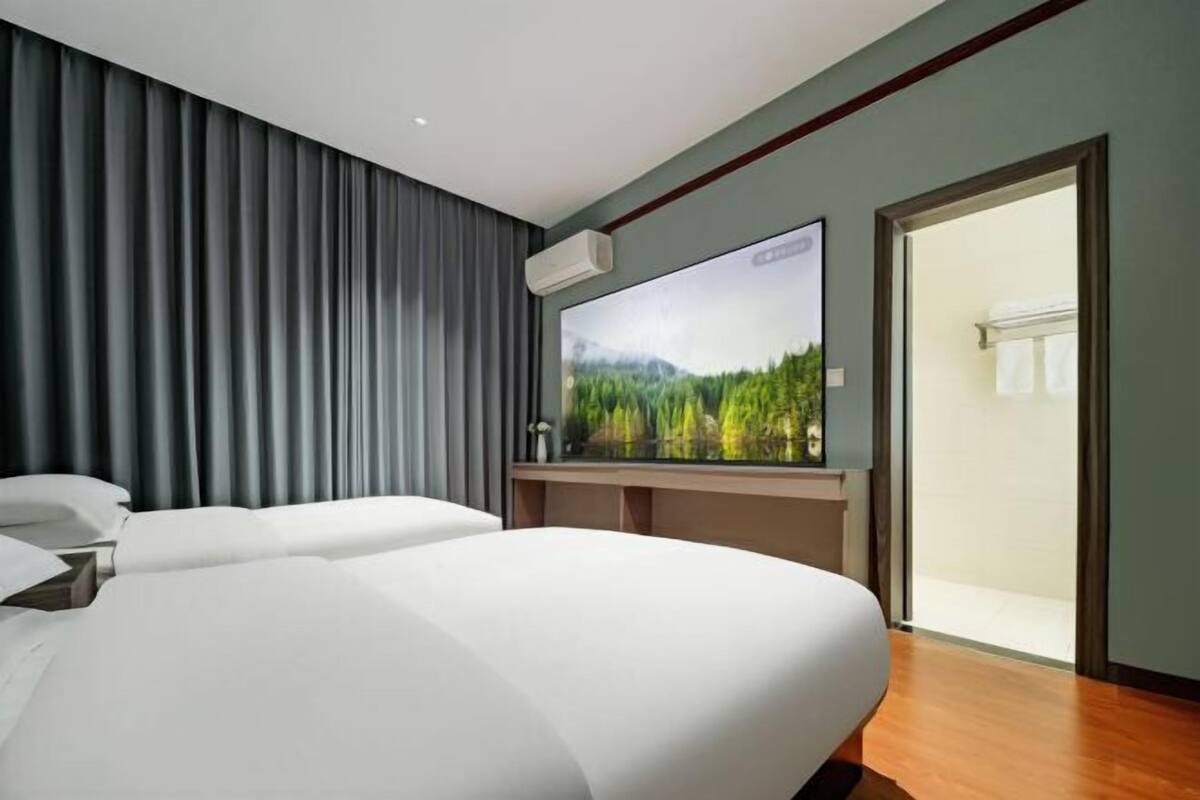 A modern hotel room with a large bed, a flat-screen TV, and a view of a forest through a window.