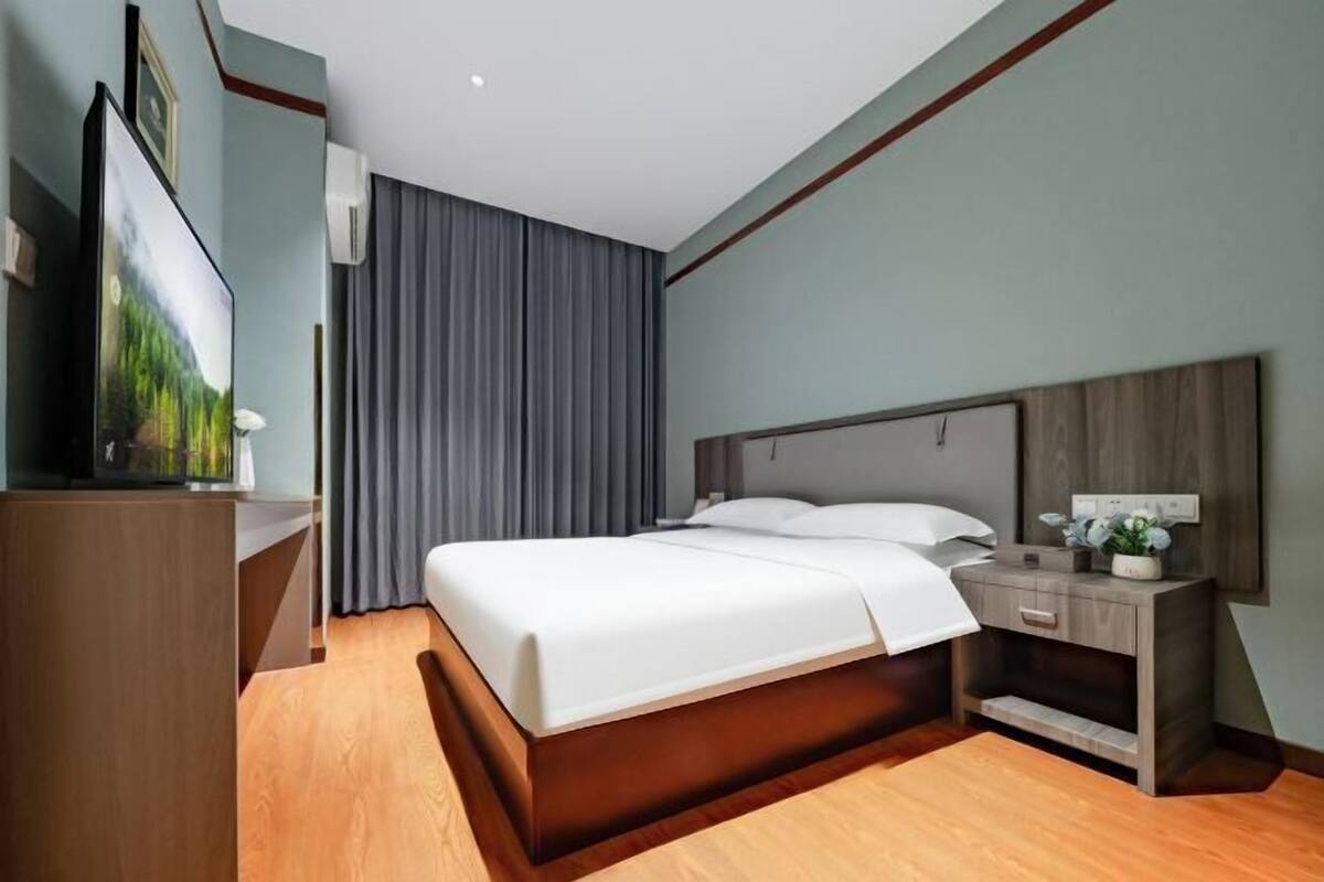 A modern hotel room with a wooden bed, a bedside table, a TV, and a large window with curtains.