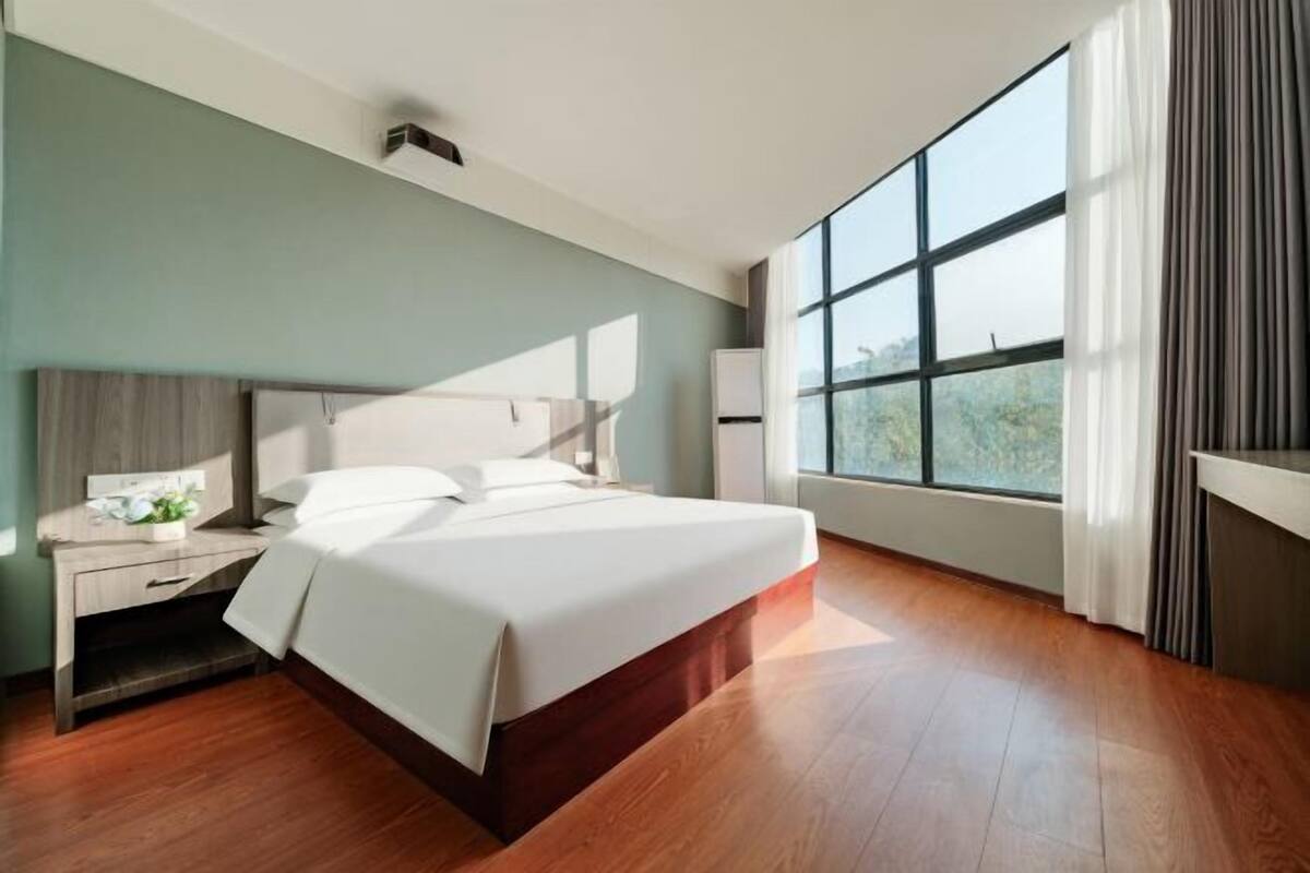 A modern hotel room with a large bed, a bedside table, a nightstand, a window with curtains, and a view of the outdoors.