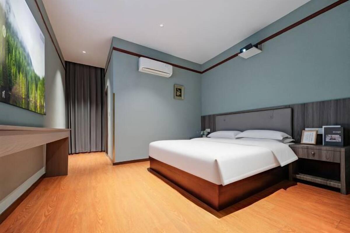 A modern hotel room with a wooden floor, a large bed with white linens, a bedside table with a lamp, and an air conditioning unit on the wall.