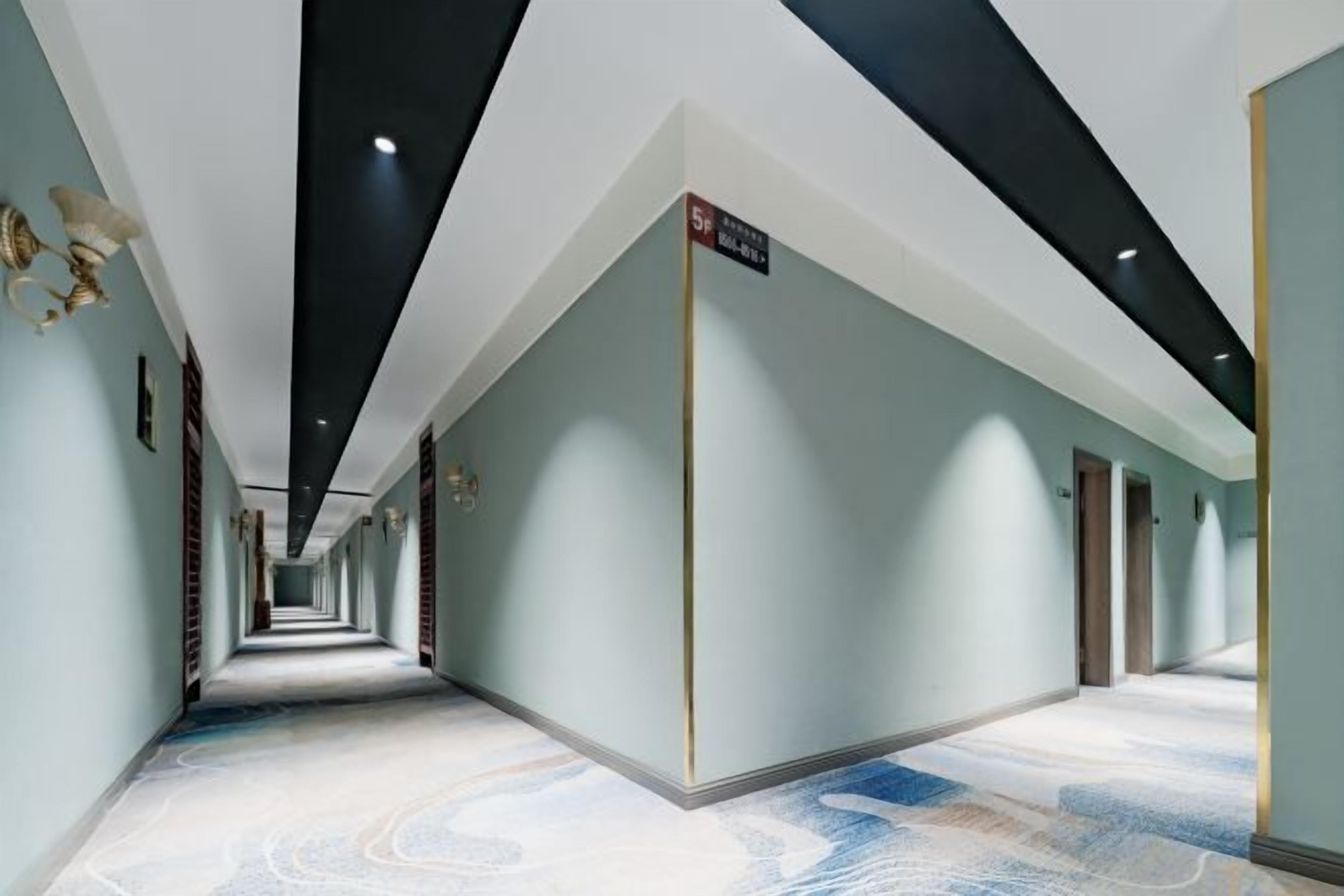 A long, narrow hallway with a patterned floor, walls with a light color, and a ceiling with black beams and recessed lighting.