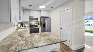 A modern kitchen with white cabinets, a granite countertop, stainless steel appliances, and a door leading to a balcony with a view of a car and greenery. - Wanna Go Back 3 Bedroom Home by RedAwning (Panama City Beach)