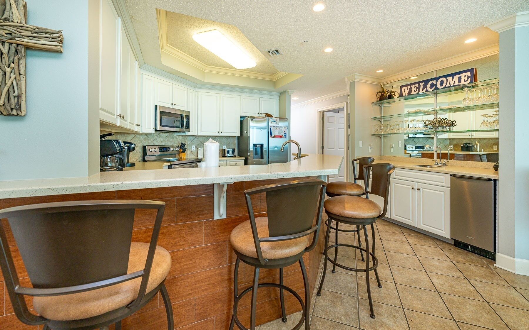 A modern kitchen with white cabinets, a stainless steel refrigerator, a microwave, and a coffee maker. There are three bar stools and a 'WELCOME' sign above the counter.