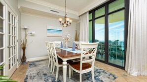 A dining area with a table, chairs, a chandelier, and a view through large windows. - Phoenix West 1706 3 Bedroom Condo (Orange Beach)