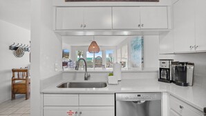 A modern kitchen with white cabinets, a stainless steel sink, a coffee maker, and a sign above stating 'Life is better at the beach.' - Bask 2 Bedroom Condo by RedAwning (Panama City Beach)