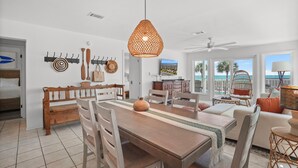 A spacious living area with a wooden dining table, a hanging light fixture, and a view of the ocean through large windows. - Bask 2 Bedroom Condo by RedAwning (Panama City Beach)