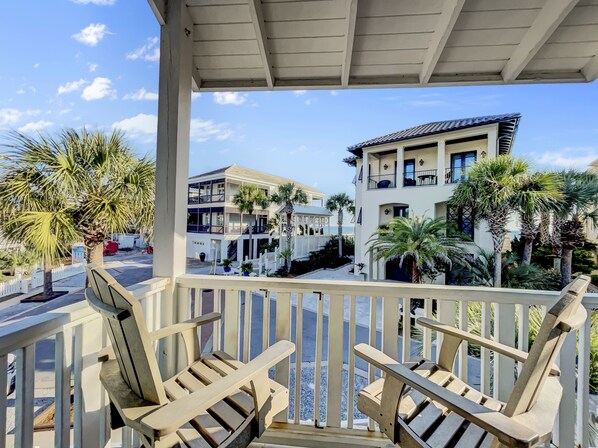 A balcony with two wooden chairs, a white railing, and a view of a multi-story building with balconies and palm trees.
