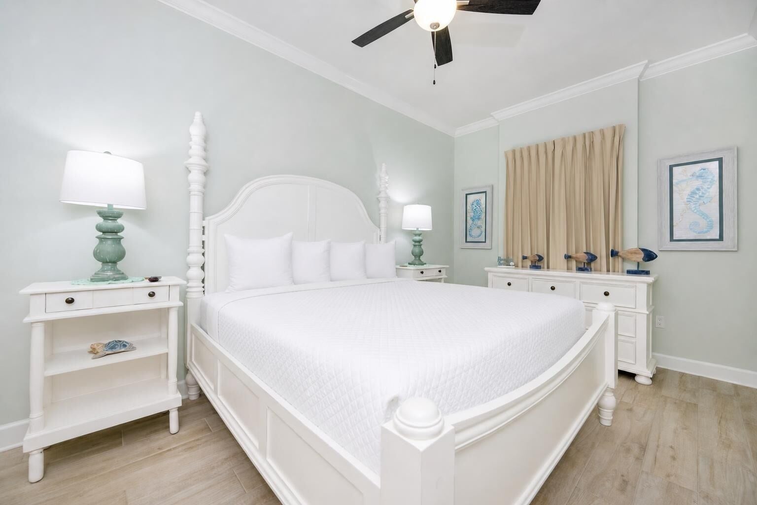 A large bed with white bedding, two bedside tables with lamps, a ceiling fan, and artwork on the walls.