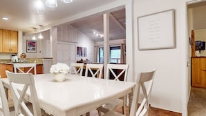 A dining area with a white table and chairs, a vase of flowers, and a kitchen in the background.