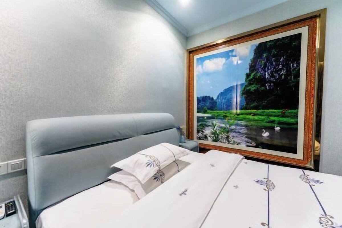 A hotel room with a bed, a framed painting of a landscape, and a window.