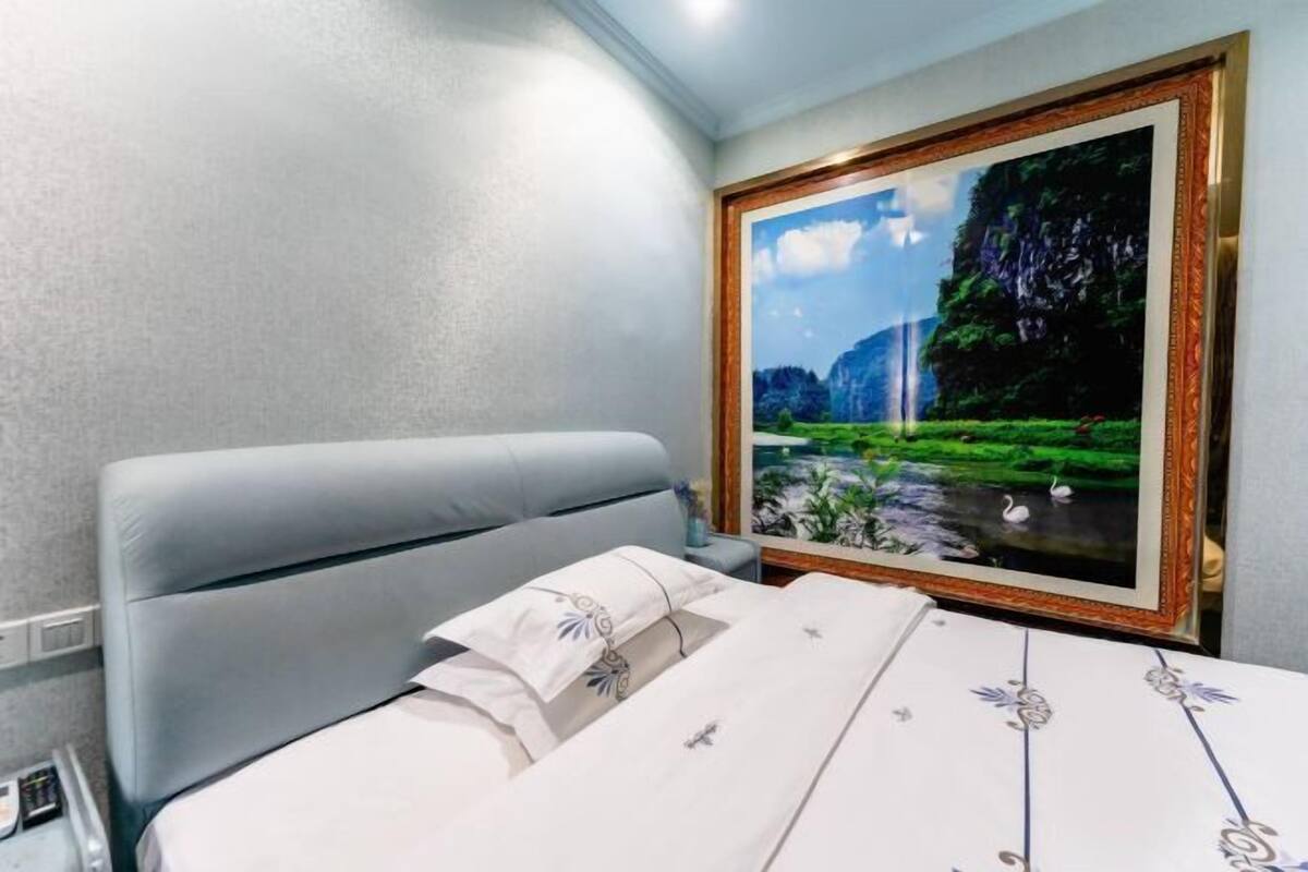 A hotel room with a bed, a headboard, a framed painting of a landscape, and a bedside table with a lamp.