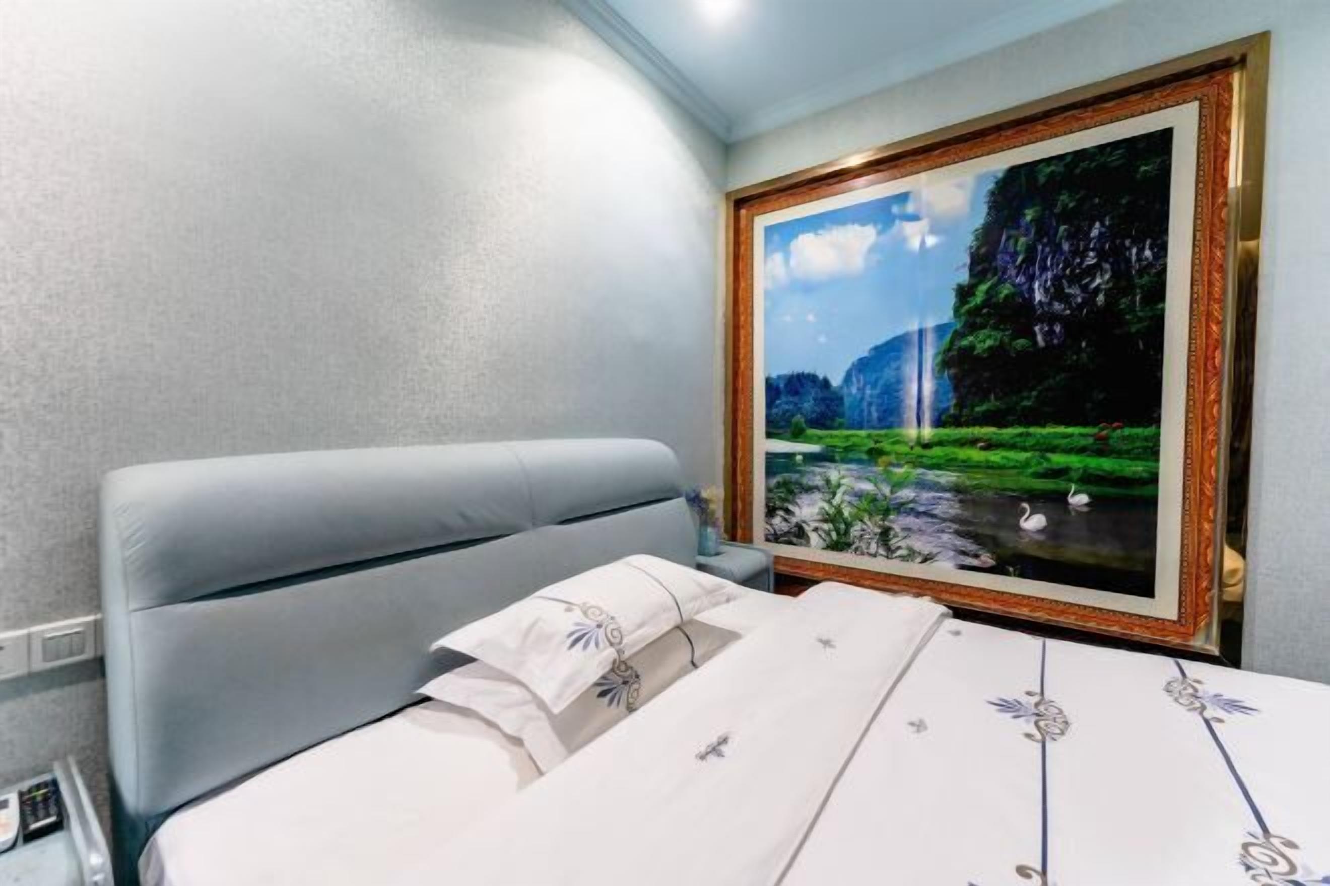 A hotel room with a bed, a painting of a landscape, and a wall-mounted light fixture.