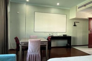 Quarto de hotel com cama, escrivaninha, cadeira, mesa e um grande ecrã em branco na parede.