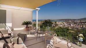 A balcony with wicker furniture, a potted plant, and a view of a coastal city. - Uefio - Luxury Suites (Monte di Procida)