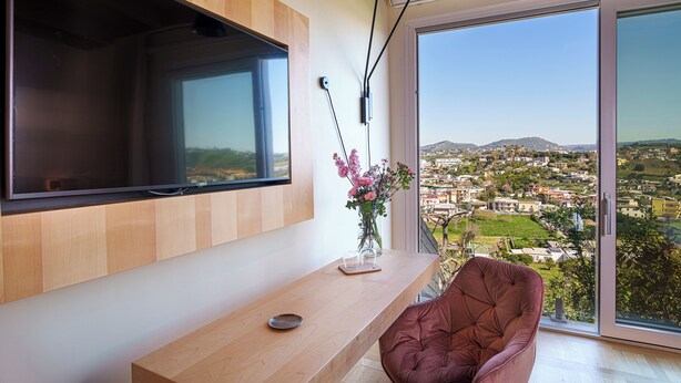 A modern living room with a large TV, a wooden table, a vase with flowers, and a view of a cityscape through a large window. - Uefio - Luxury Suites (Monte di Procida)