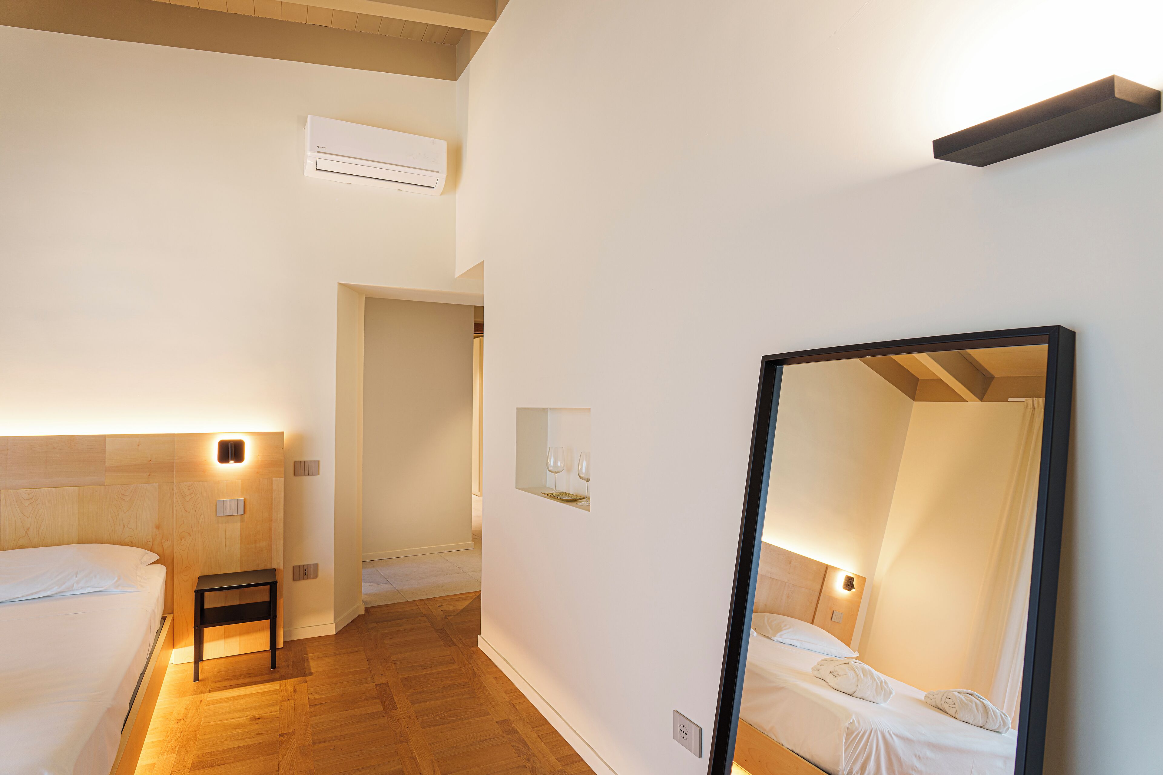 Ein Hotelzimmer mit Holzboden, einem Bett mit weißen Leinentüchern, einem Nachttisch, einem Spiegel und einer wandmontierten Klimaanlage.