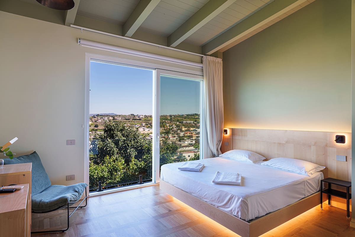 A modern bedroom with a large bed, a desk, and a view of a cityscape through a sliding glass door.