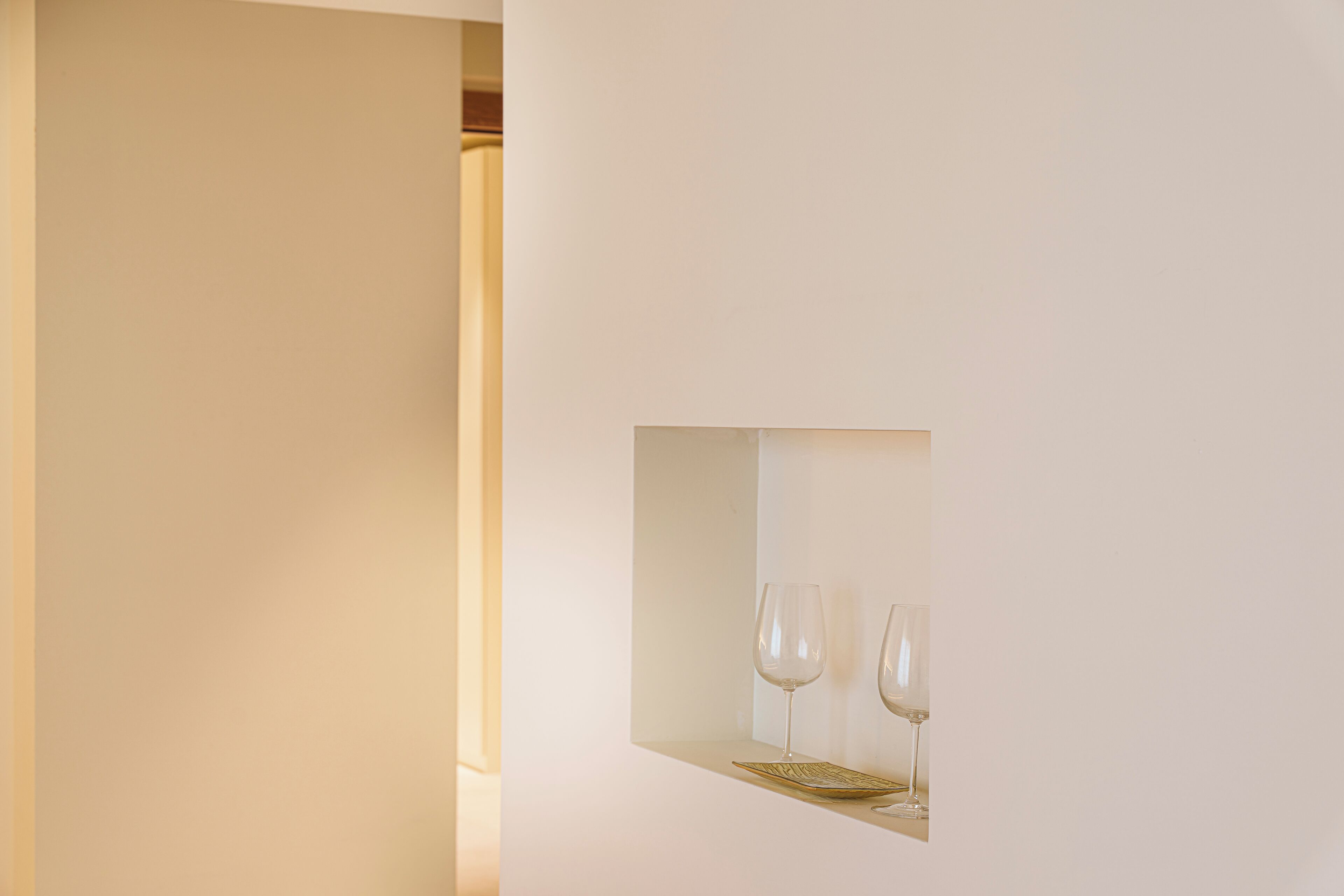 Un interior minimalista con pared blanca y un pequeño nicho que contiene dos copas de vino y una bandeja dorada.