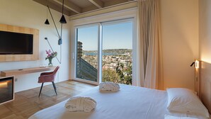 A modern hotel room with a large bed, a TV mounted on the wall, a desk with a chair, and a view of the city through a large window. - Uefio - Luxury Suites (Monte di Procida)