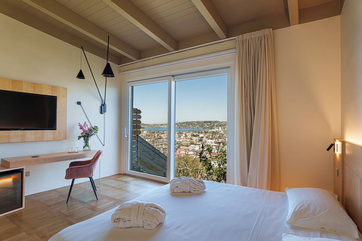 A modern hotel room with a large bed, a TV mounted on the wall, a desk with a chair, and a view of the city through a large window.