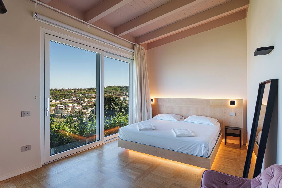 A bedroom with a large sliding glass door, a bed with white linens, and a view of a cityscape.