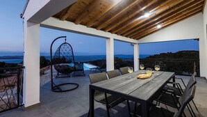 An outdoor patio with a wooden table, chairs, a hanging chair, and a pool view.