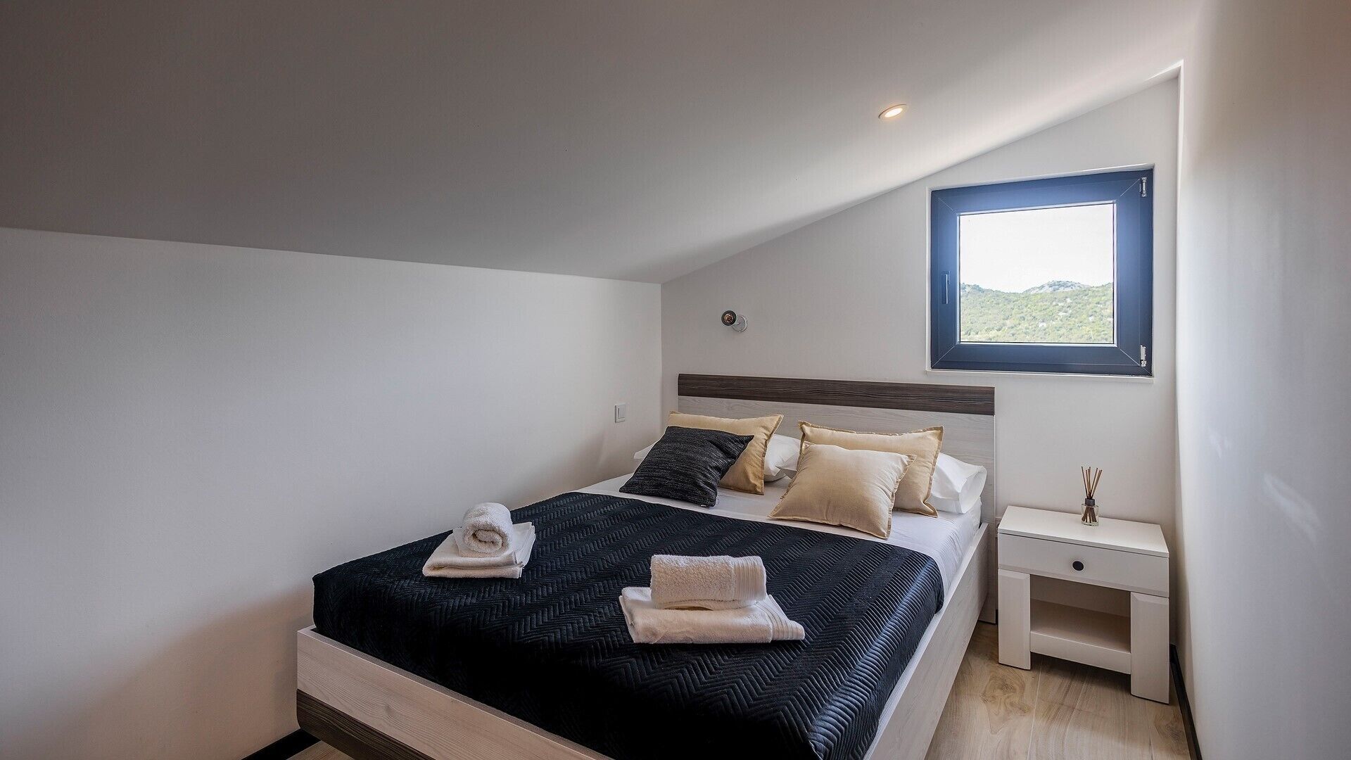A modern bedroom with a large bed, a nightstand, and a window with a view of greenery.