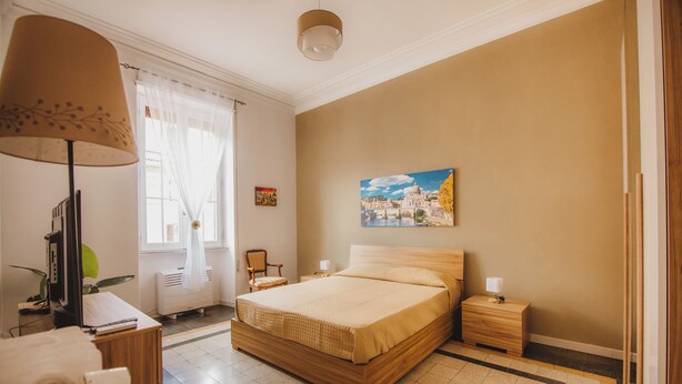 A bedroom with a bed, bedside tables, a desk with a lamp, a chair, and a window with curtains. - Grazioli Vatican Home (Roma)
