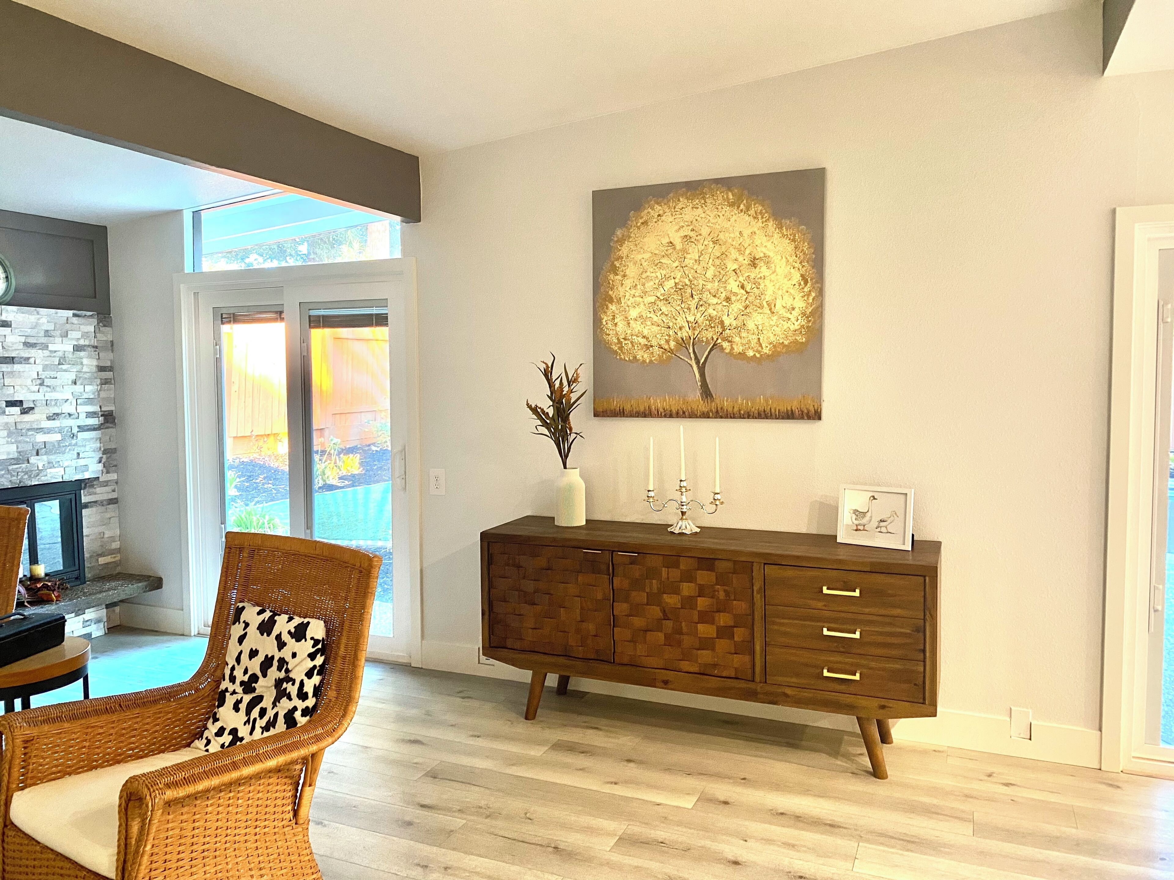 A living room with a wicker chair, a wooden cabinet, and a painting of a tree.
