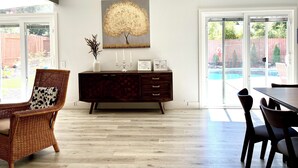 Un comedor con una mesa y sillas de madera oscura, una silla de mimbre con cojín estampado con vaca, un mueble de madera oscura con un jarrón y fotografías enmarcadas, y un ventanal con vista a un patio trasero.