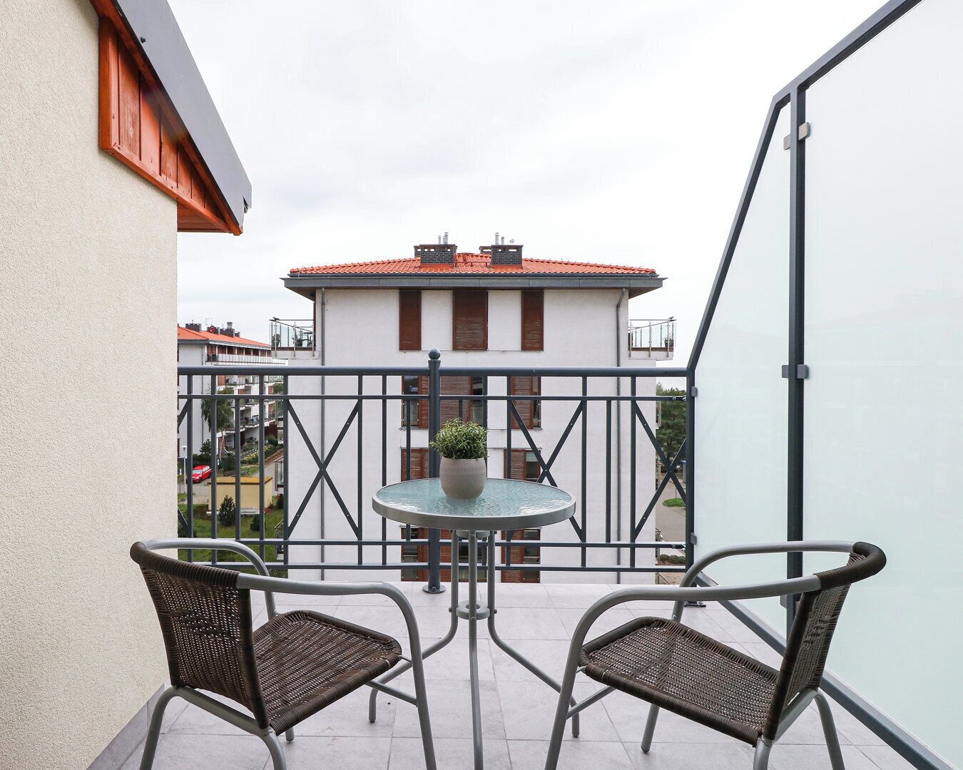 Un balcone con due sedie in vimini e un piccolo tavolo rotondo, con vista su una zona residenziale dai tetti in tegole rosse.