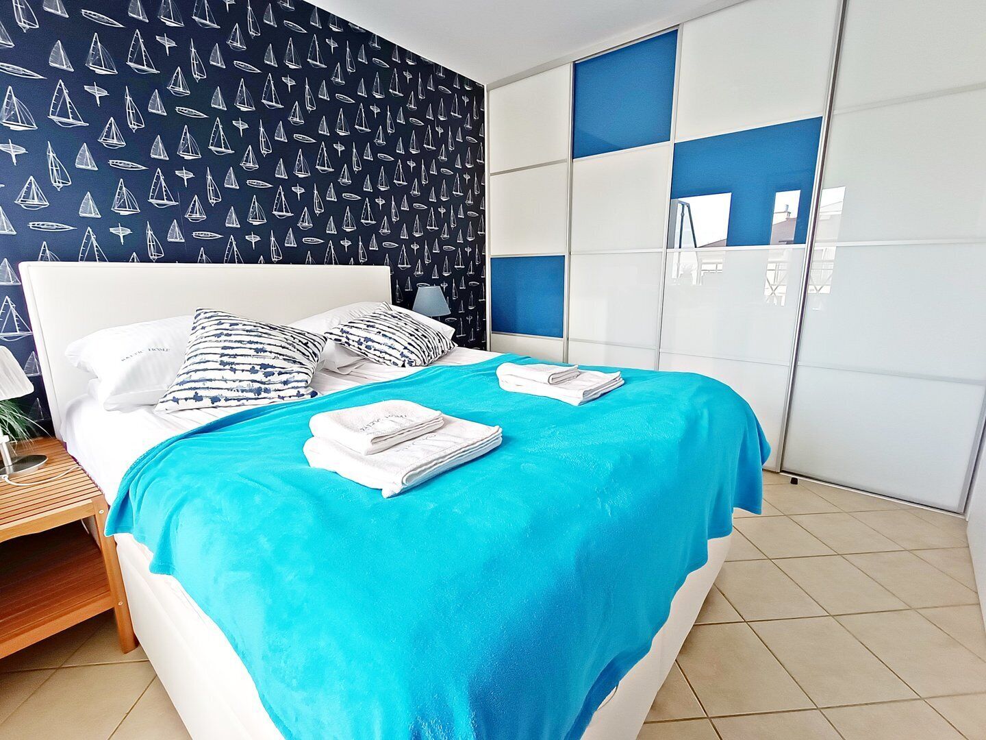 A neatly made bed with a blue bedspread and white pillows, a wooden nightstand with a lamp, and a wardrobe with blue panels.