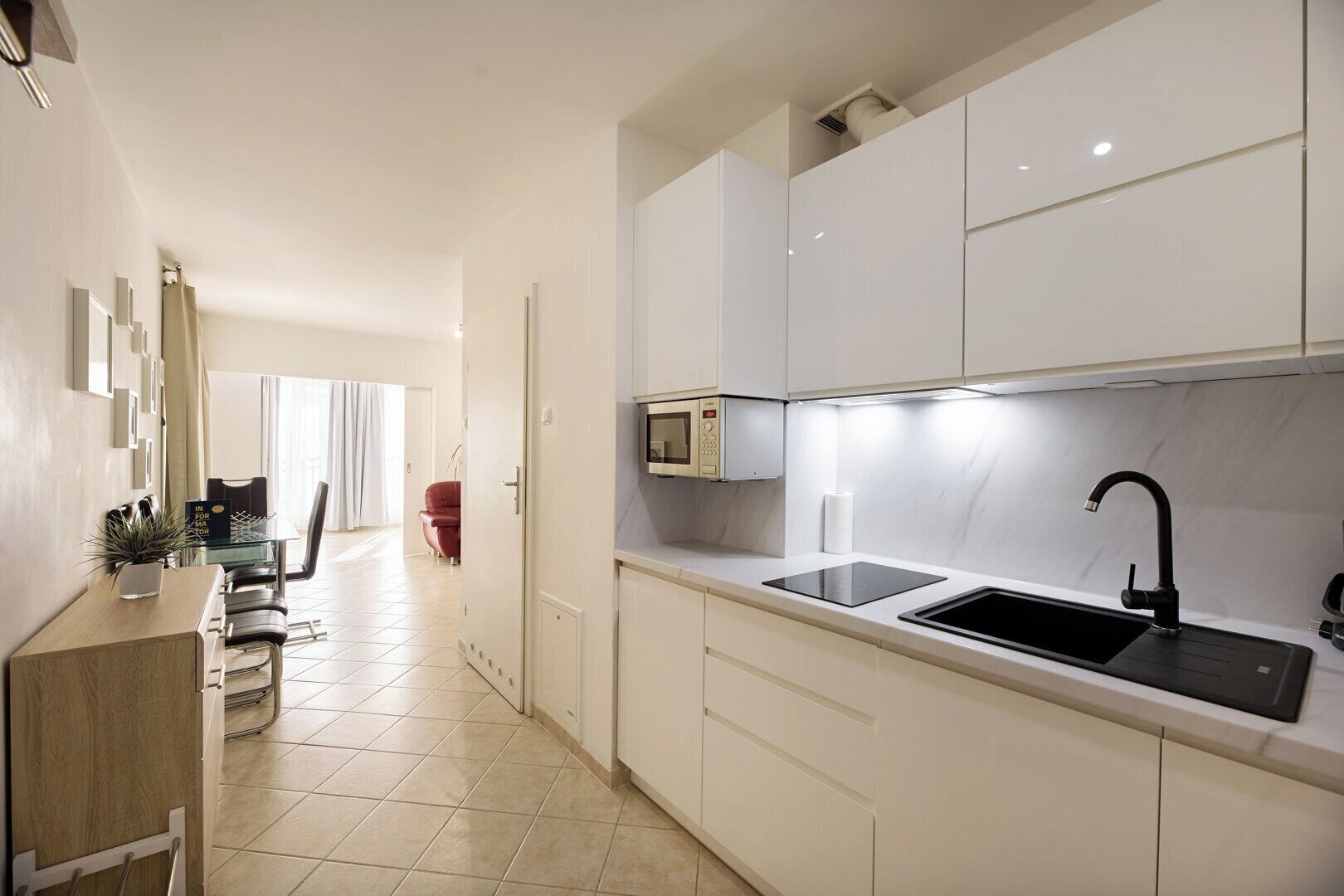 Cocina moderna con armarios blancos, un lavamanos negro y un microondas. Hay un comedor con una mesa y sillas de vidrio, y una silla roja al fondo.