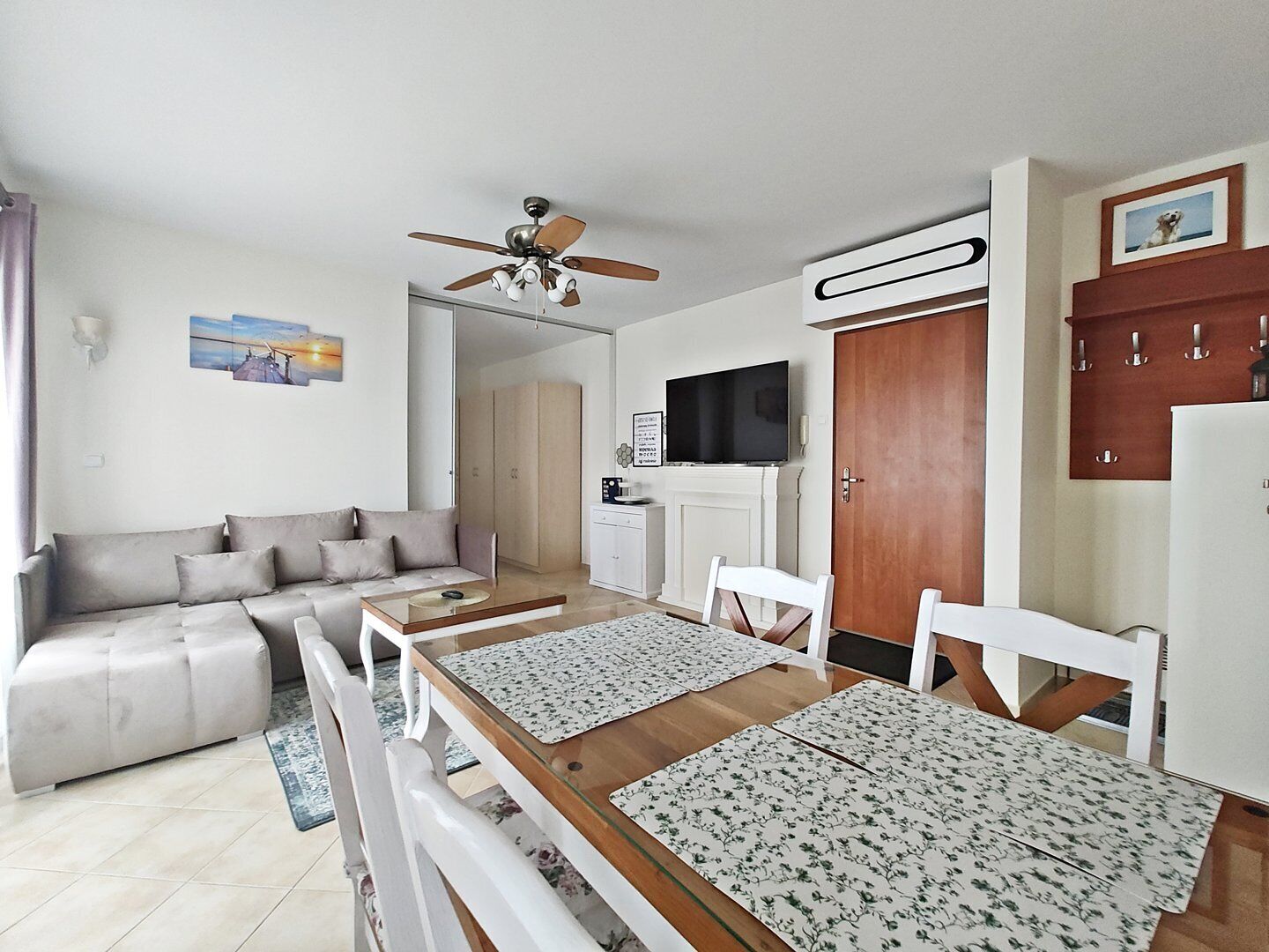 Ein modernes Wohnzimmer mit einer Couch, einem Holzes Esstisch mit Platzdeckchen, einem Deckenventilator, einem Fernseher und einem Kleiderschrank.