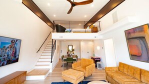 A spacious living area with a high ceiling fan, a staircase, a dining area, and a large painting on the wall.